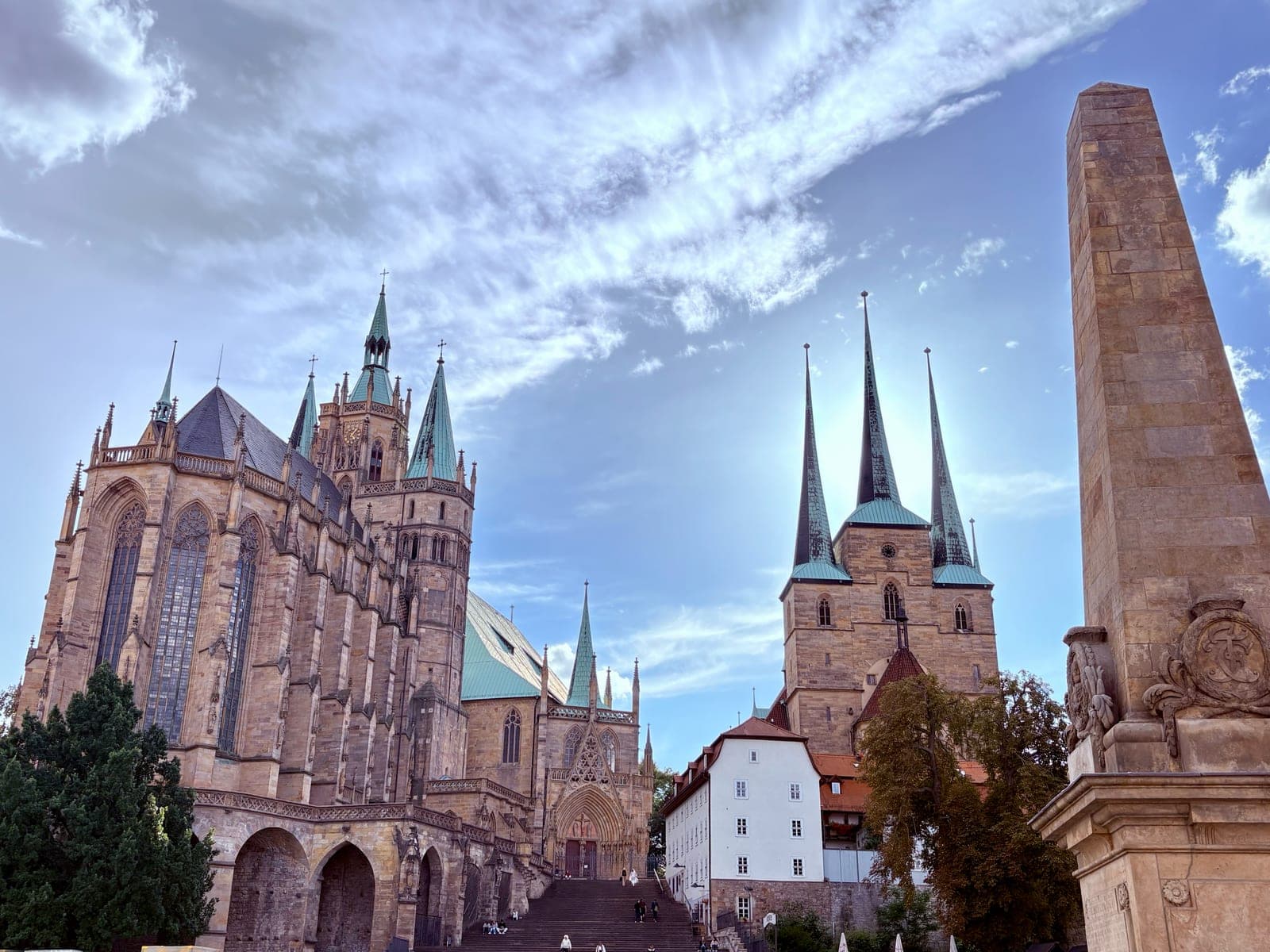 Erfurt's Domplatz under a streaked blue sky: the Gothic cathedral with its steps on the left, the three spires of St. Severus in the middle and a sandstone obelisk at the right edge